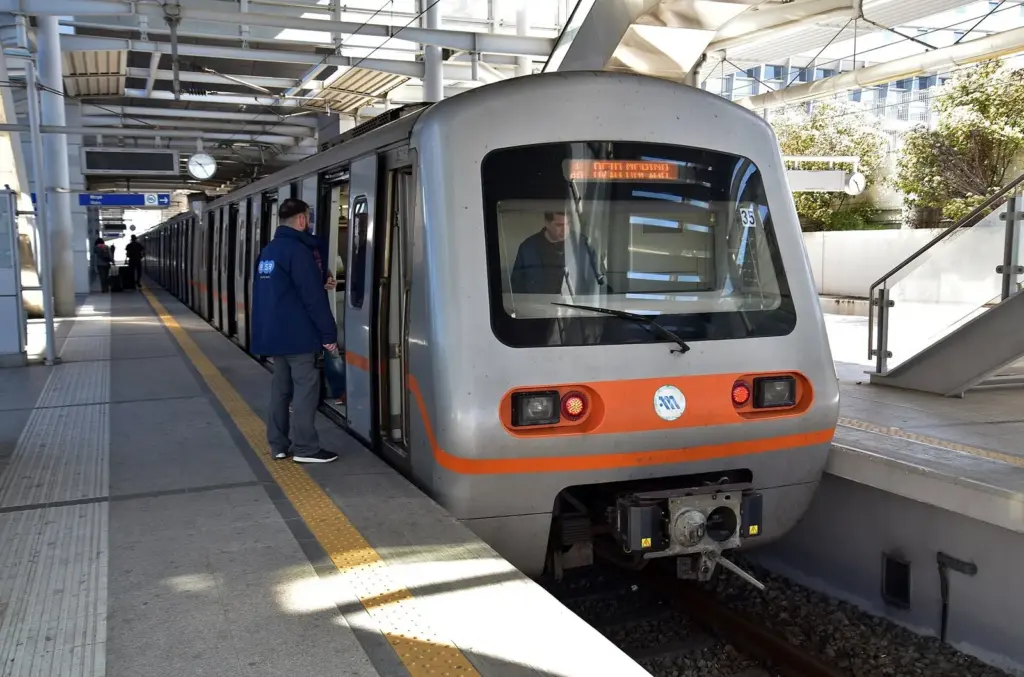 A silver Line 3 metro train with an orange stripe at the airport station platform, the direct rail link from Athens airport to Piraeus port