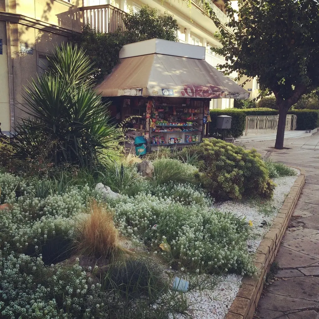 A periptero kiosk with a canvas awning behind a planted verge on a pavement in Ampelokipoi, Athens