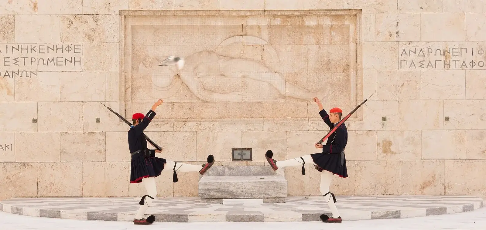 Two Evzones in navy winter uniform kicking in unison before the carved relief of the Tomb of the Unknown Soldier