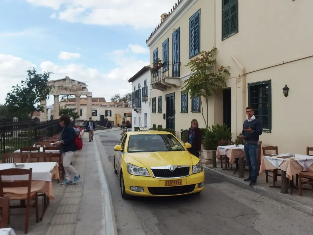 A yellow taxi beside taverna tables on a narrow street, with ancient columns at its far end