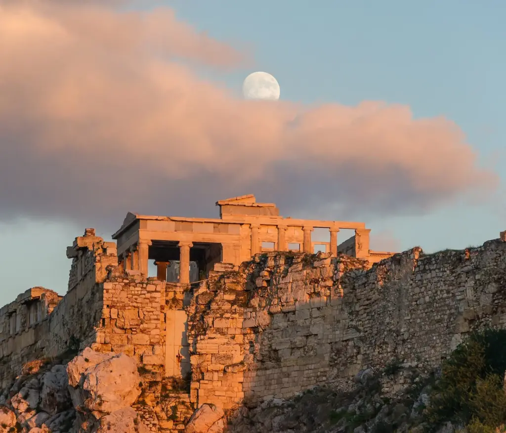 The Erechtheion and the Acropolis wall lit orange by the setting sun, with the moon above