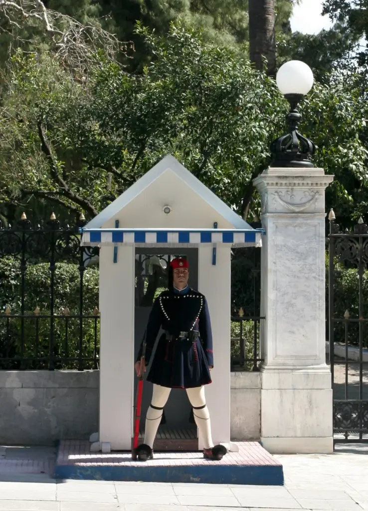 An Evzone in navy uniform standing in a blue-and-white sentry box beside the gate of the Presidential Mansion