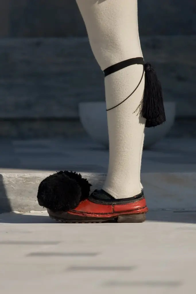 Close-up of an Evzone's red leather tsarouhi with its black pompom and a tasselled garter on the white stocking