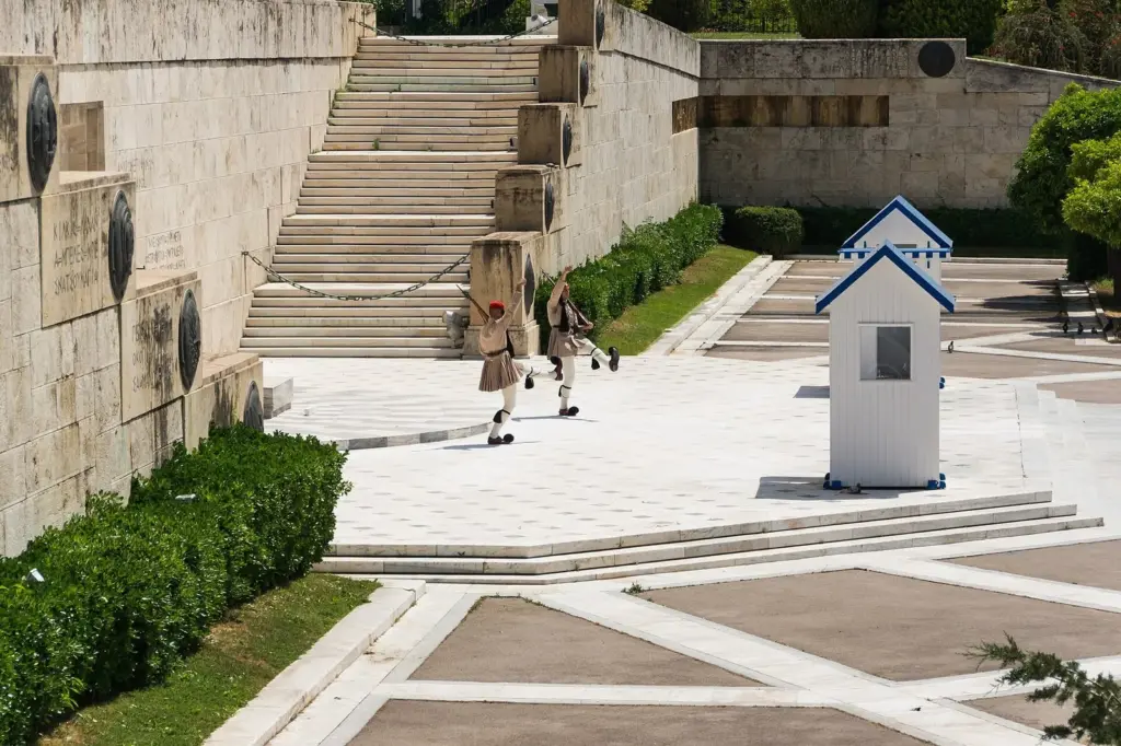 Two Evzones in khaki summer uniform crossing the paved forecourt past a sentry box below the Parliament