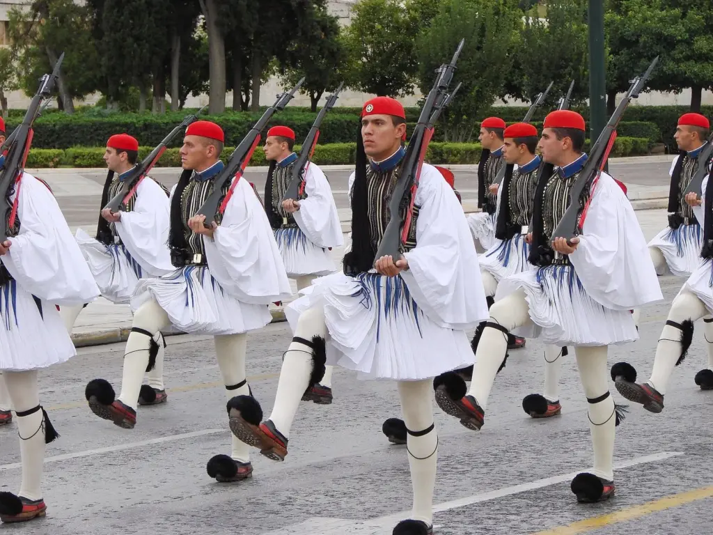 A column of Evzones in white fustanellas and dark embroidered waistcoats marching with rifles shouldered