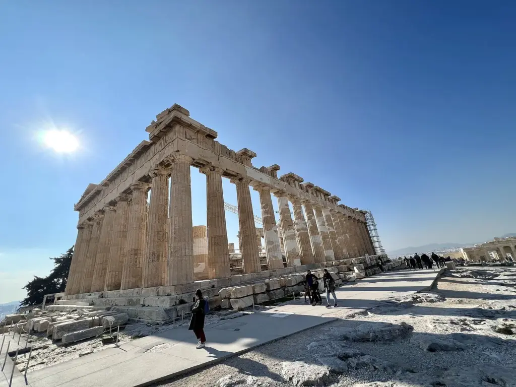 The Parthenon's long colonnade under a bright sun, with a few visitors on the paved path beside it