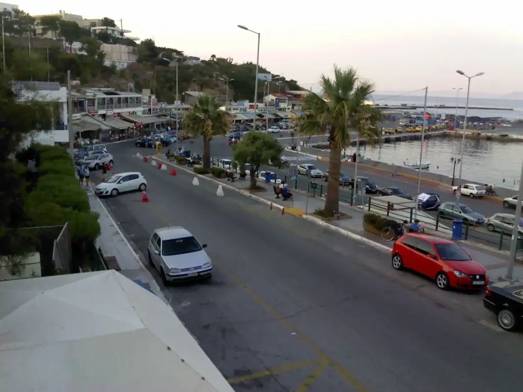 Harbour-front road in Rafina with palm trees, parked cars and café awnings, the port beyond