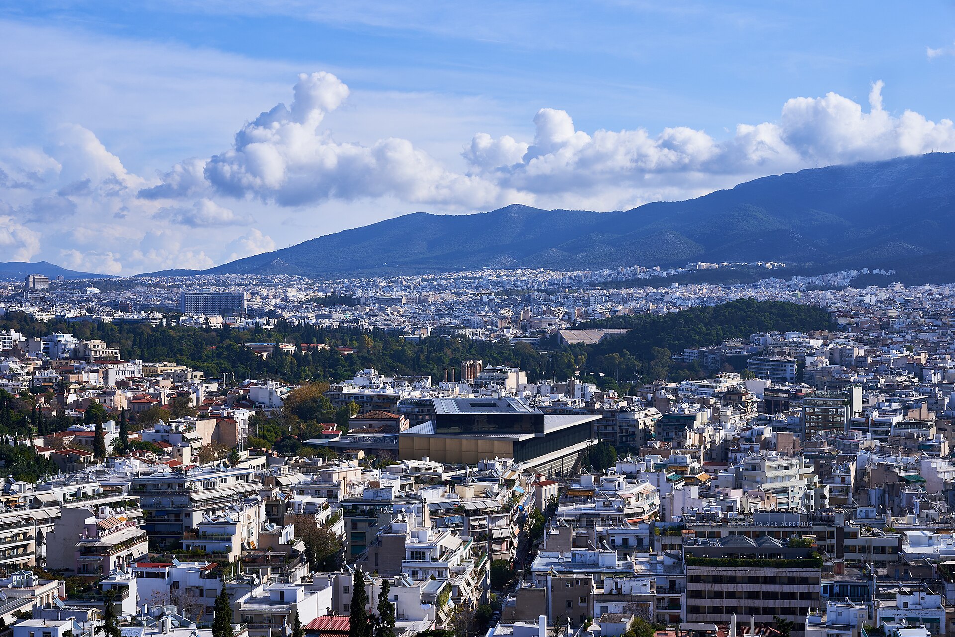 The Acropolis Museum stands among the buildings of Athens, its dark glass upper floor visible below the distant mountains.