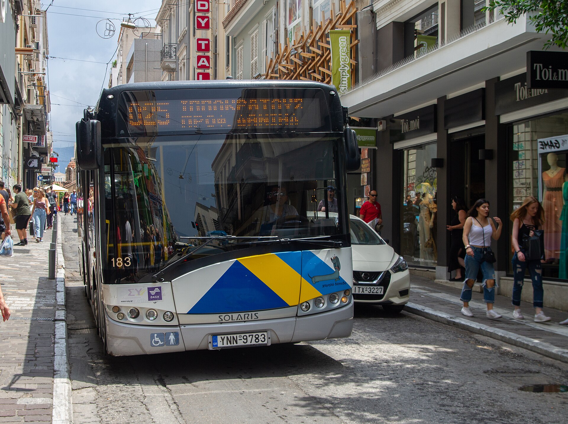 An Athens city bus faces the viewer on a narrow shopping street with pedestrians walking along both pavements.