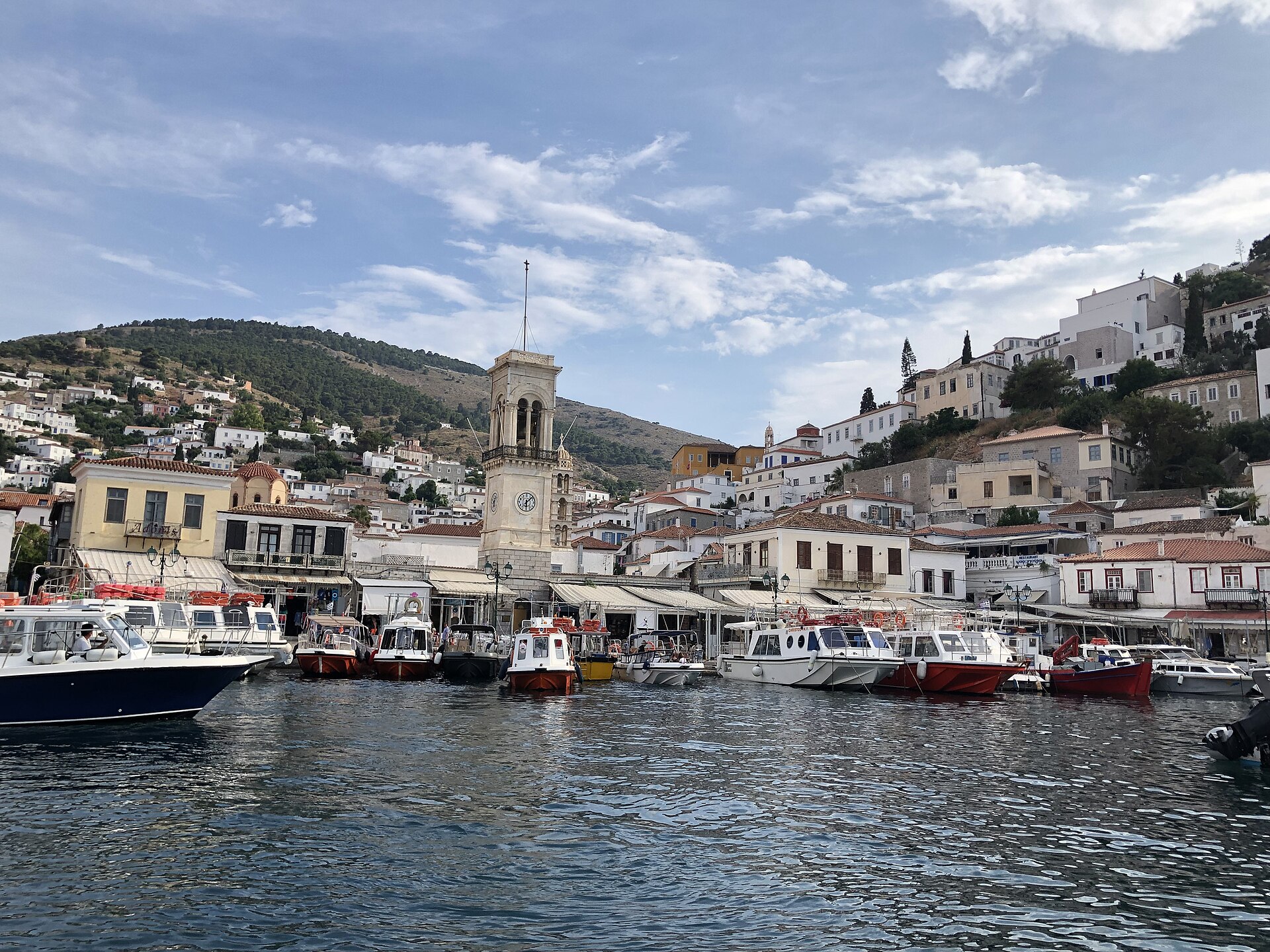 Boats line Hydra town harbour beneath a clock tower, while stone mansions and whitewashed houses rise on the surrounding slopes.