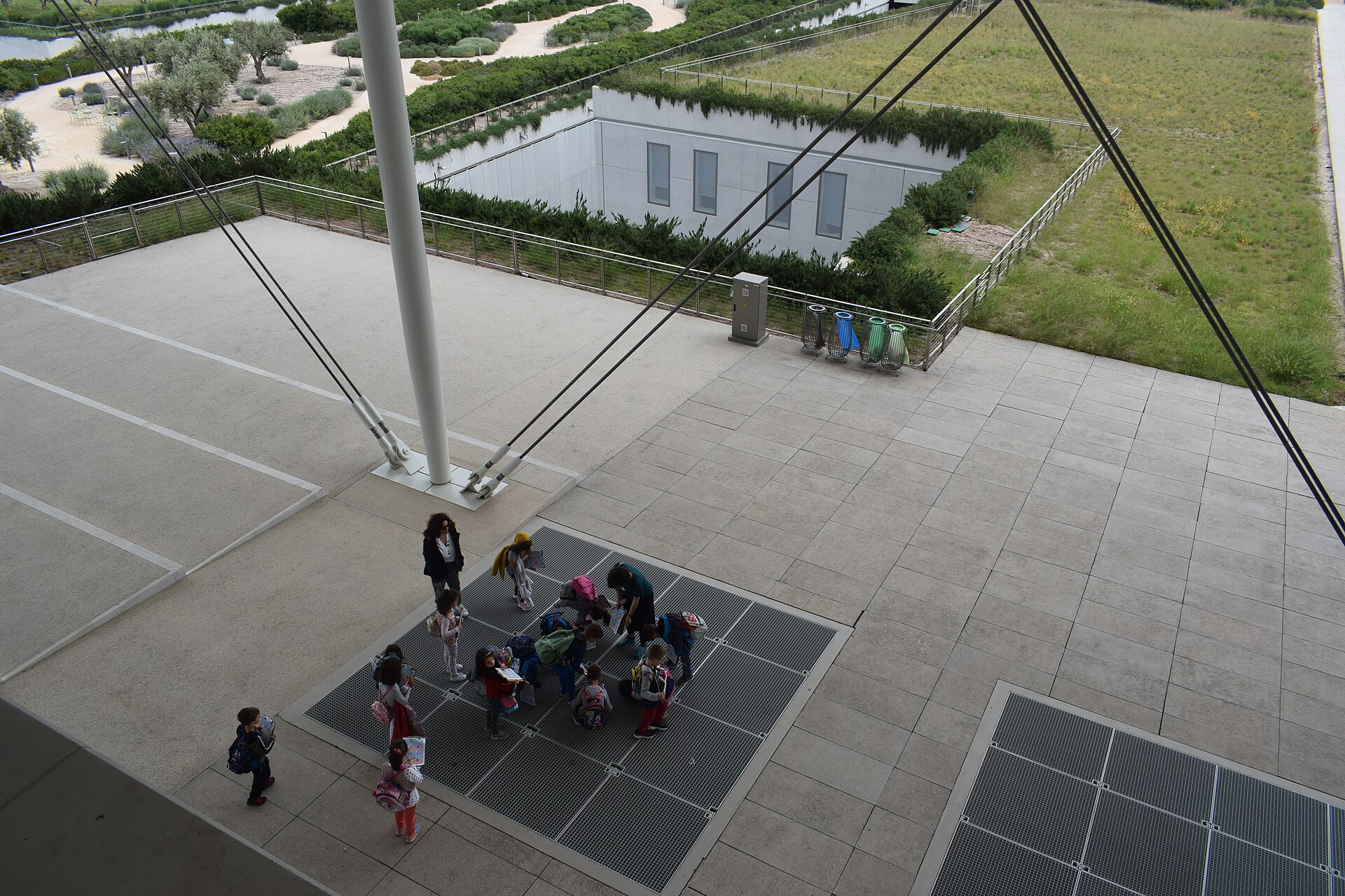 Children and adults gather beneath the SNFCC canopy on a terrace beside planted roofs and garden paths in Athens.