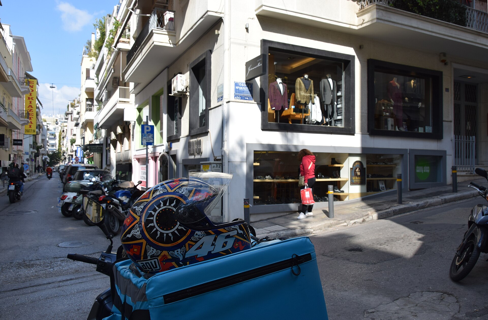 Clothing displays fill corner windows on Tsakalof Street in Kolonaki, Athens, beside pedestrians and parked scooters.