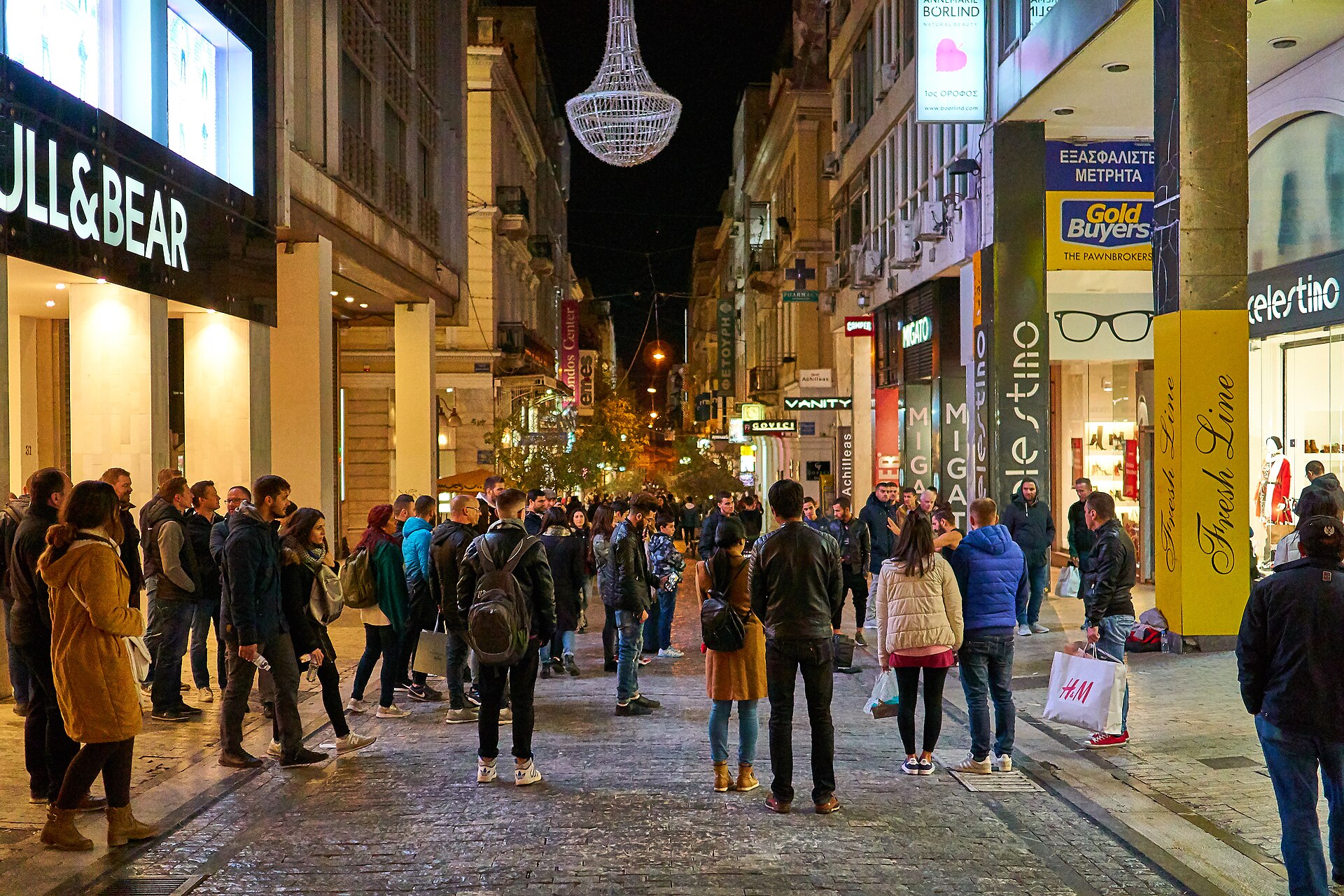 Evening shoppers walk along pedestrian Ermou Street in Athens between illuminated storefronts and beneath a hanging chandelier decoration.