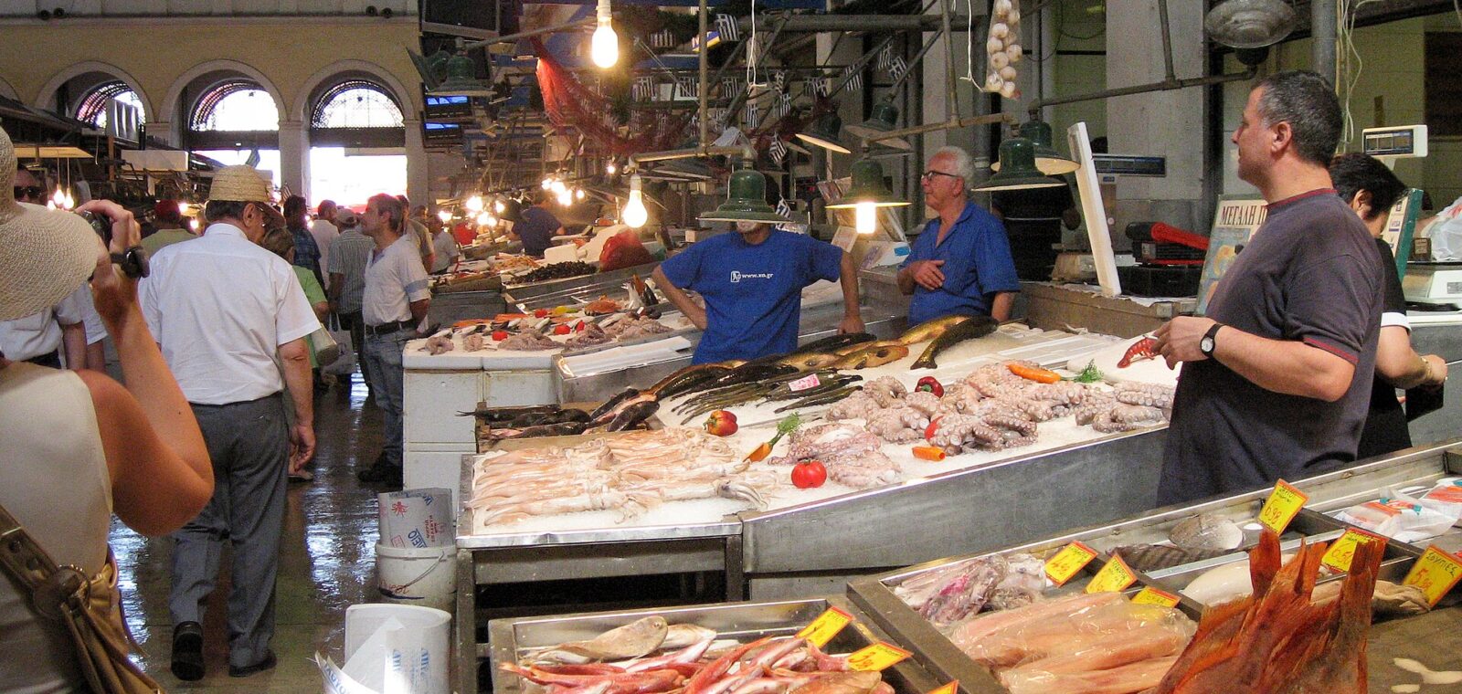 Fishmongers stand behind seafood counters beneath high windows inside Athens Central Market, while shoppers walk along the aisle.