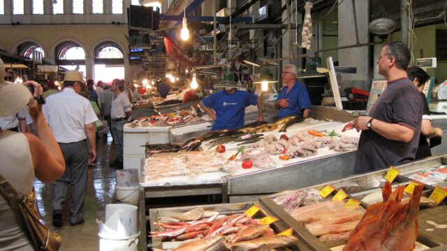Fishmongers stand behind seafood counters beneath high windows inside Athens Central Market, while shoppers walk along the aisle.