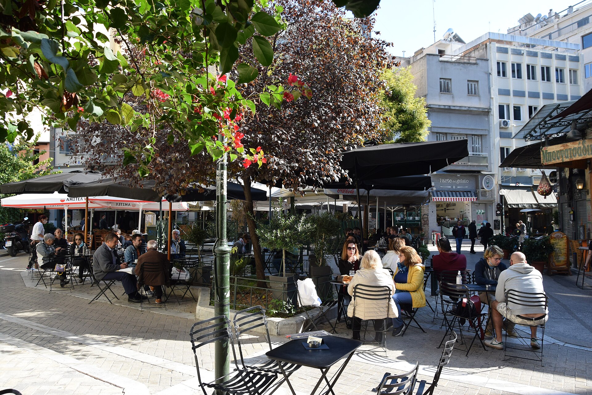 Groups sit at outdoor tables beneath parasols and leafy trees on Iroon Square in Psyrri, Athens.