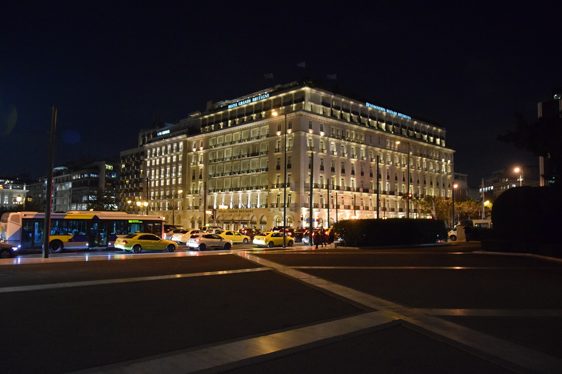 Hotel Grande Bretagne in central Athens is illuminated at night above a street filled with taxis and a city bus.