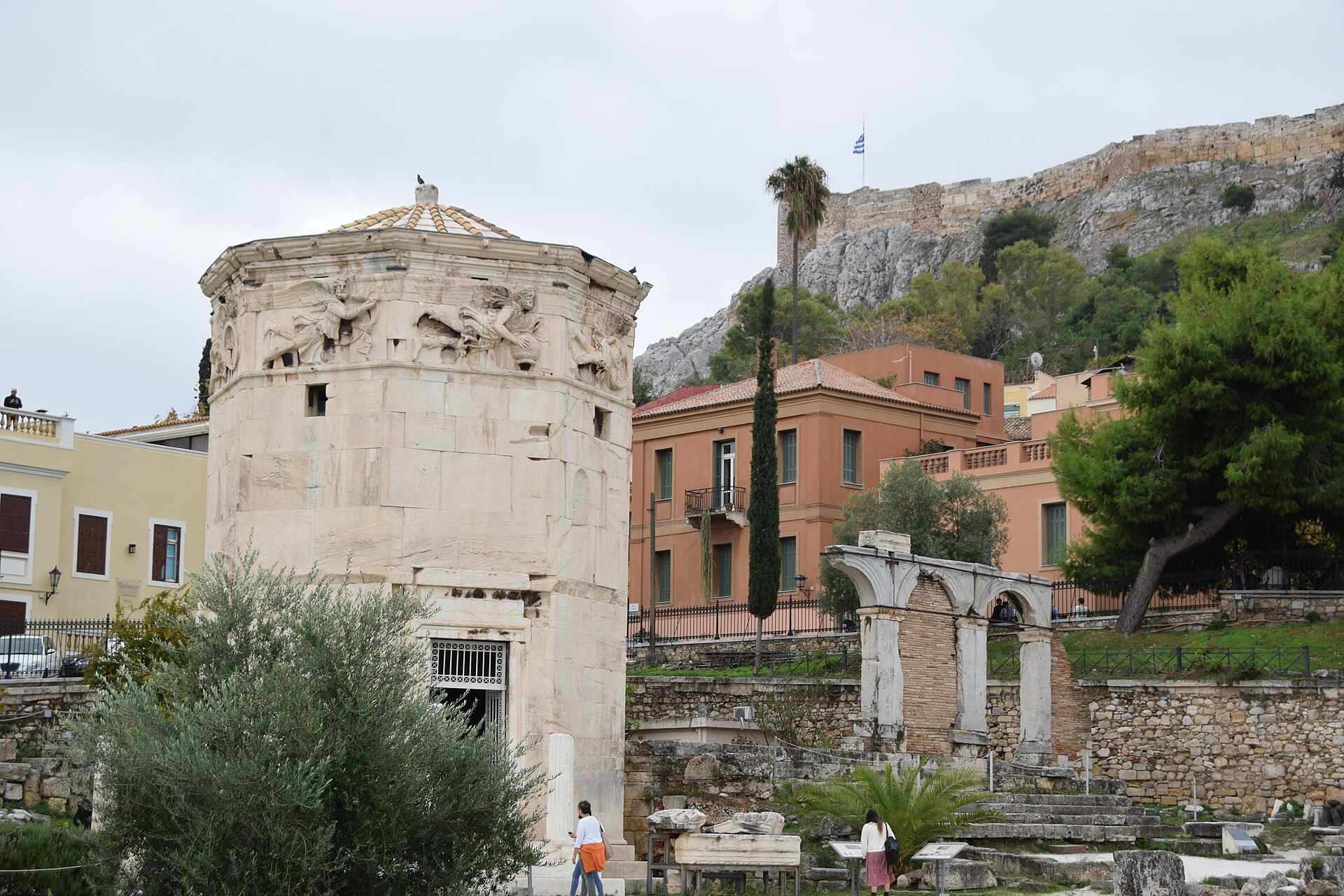 The marble Tower of the Winds rises beside arched ruins in Athens, with carved figures beneath its roof.