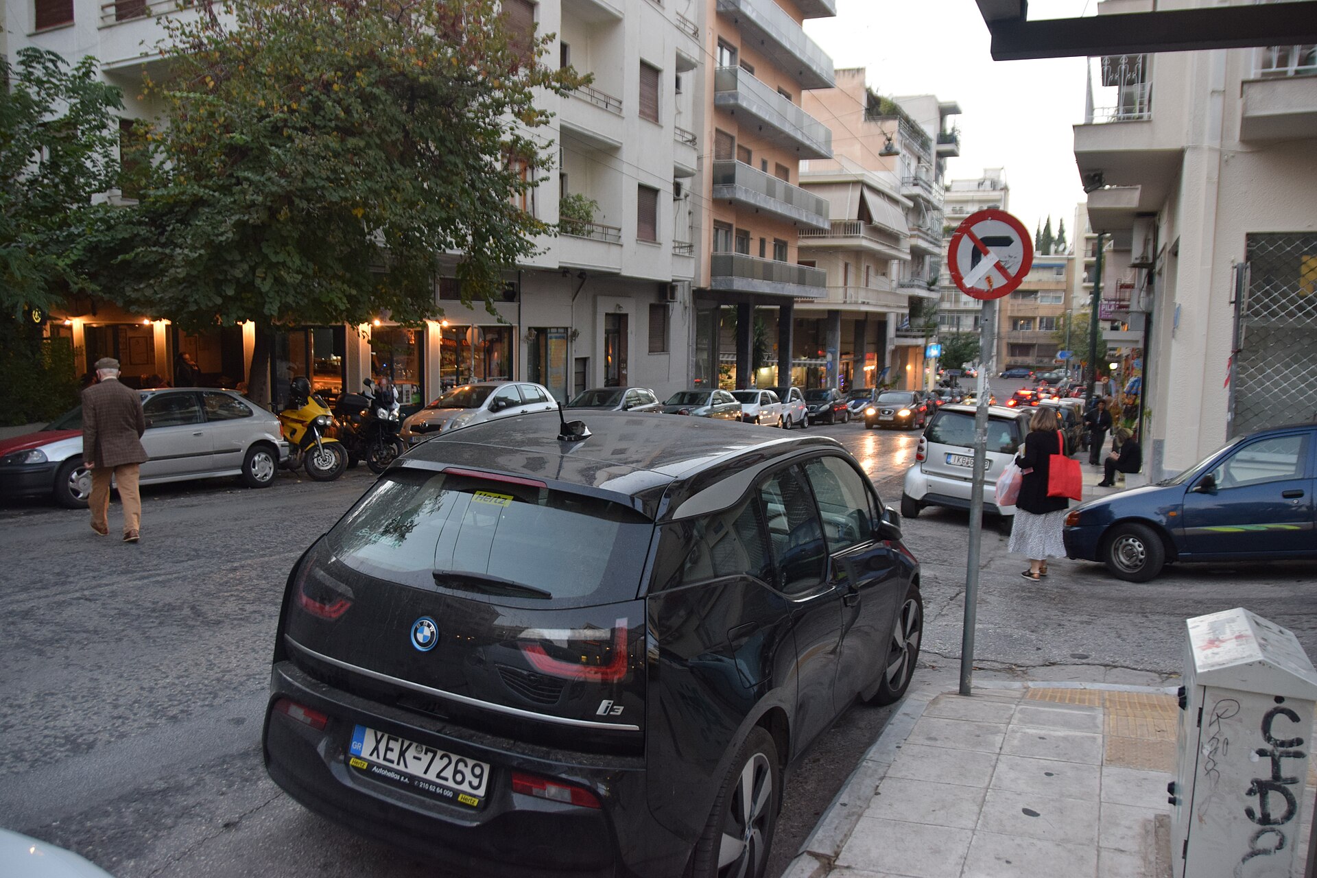 Pedestrians pass parked cars and tree-shaded ground-floor premises beneath apartment balconies on Eratosthenous Street in Pangrati, Athens.