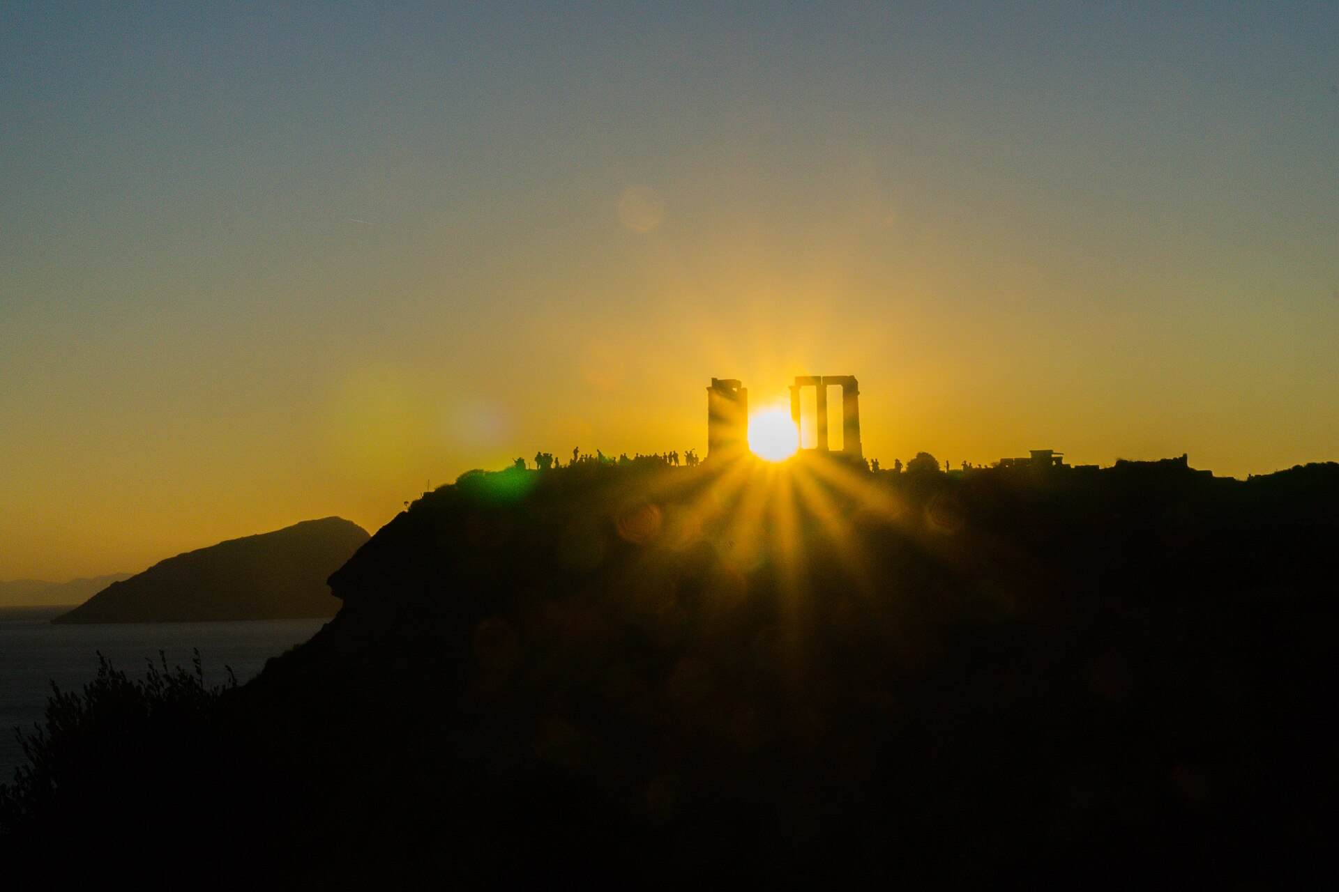 The setting sun shines through the Temple of Poseidon at Cape Sounion above a sea cliff lined with visitors.