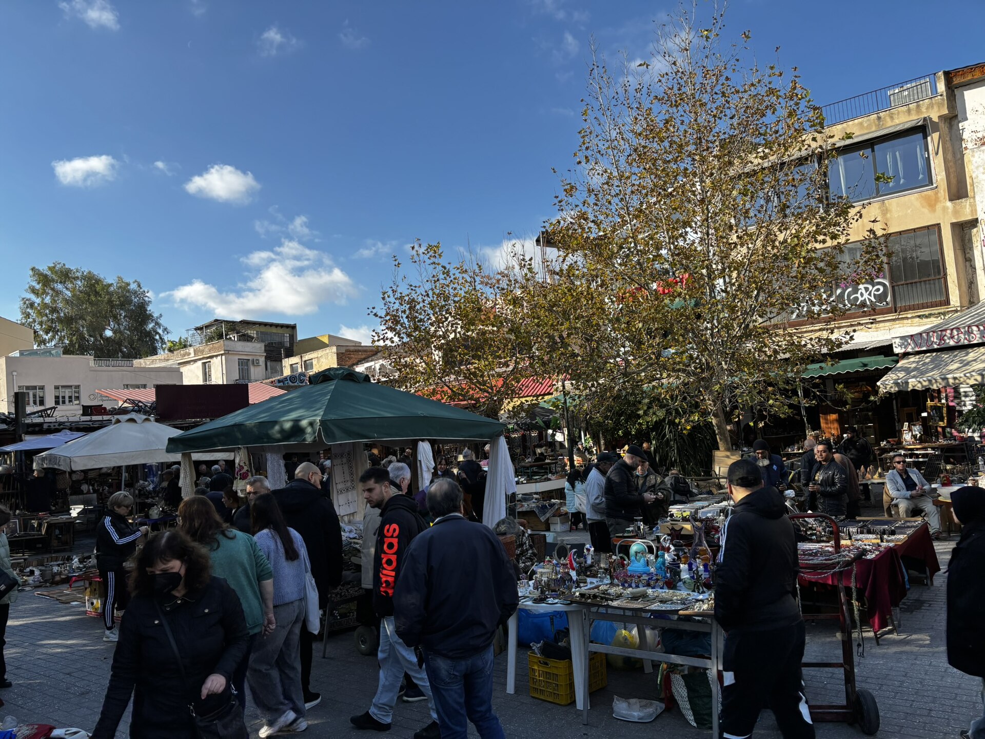Shoppers browse tables of glassware and bric-a-brac under umbrellas at the Monastiraki flea market in Athens.