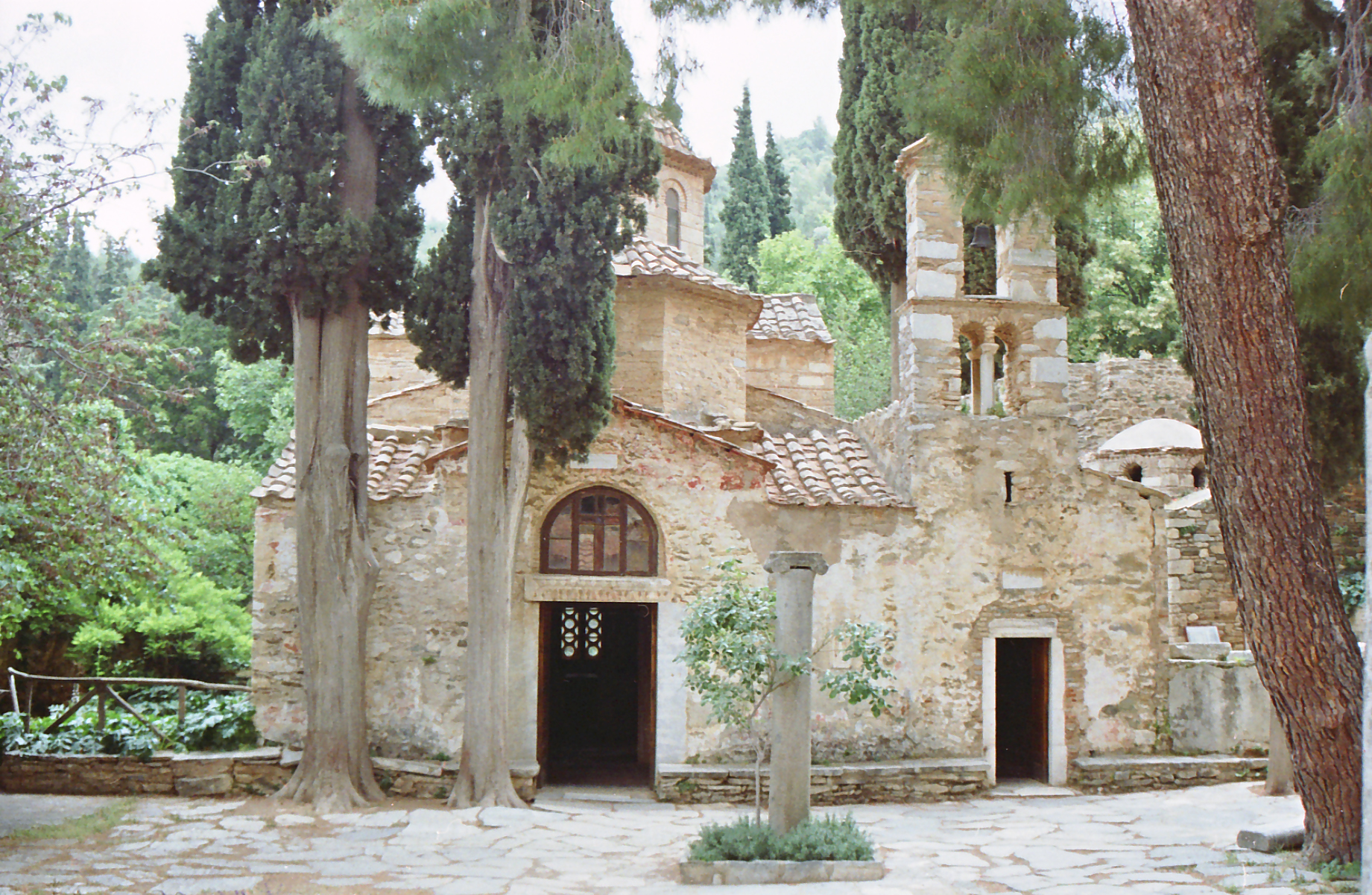 The stone church at Kaisariani Monastery stands in a tree-shaded courtyard, with an arched doorway and a narrow bell tower.