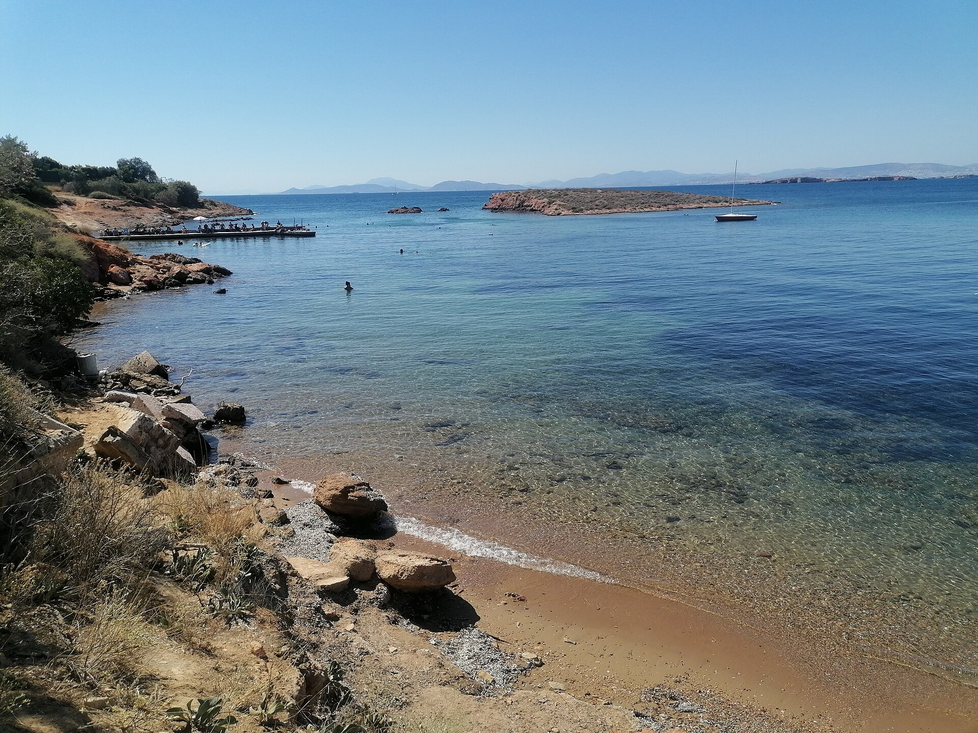 Swimmers enter clear water beside a small Kavouri beach, with rocky islets and a moored sailboat across the bay.