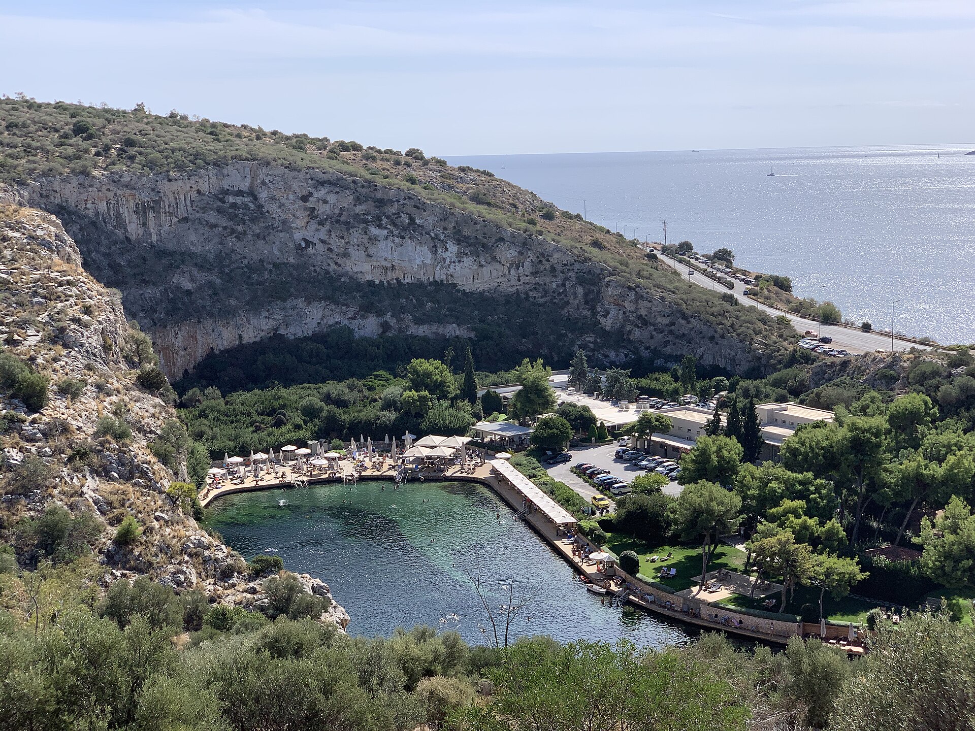 Swimmers and loungers surround Lake Vouliagmeni beneath a limestone cliff, with the coastal road and sea visible beyond.
