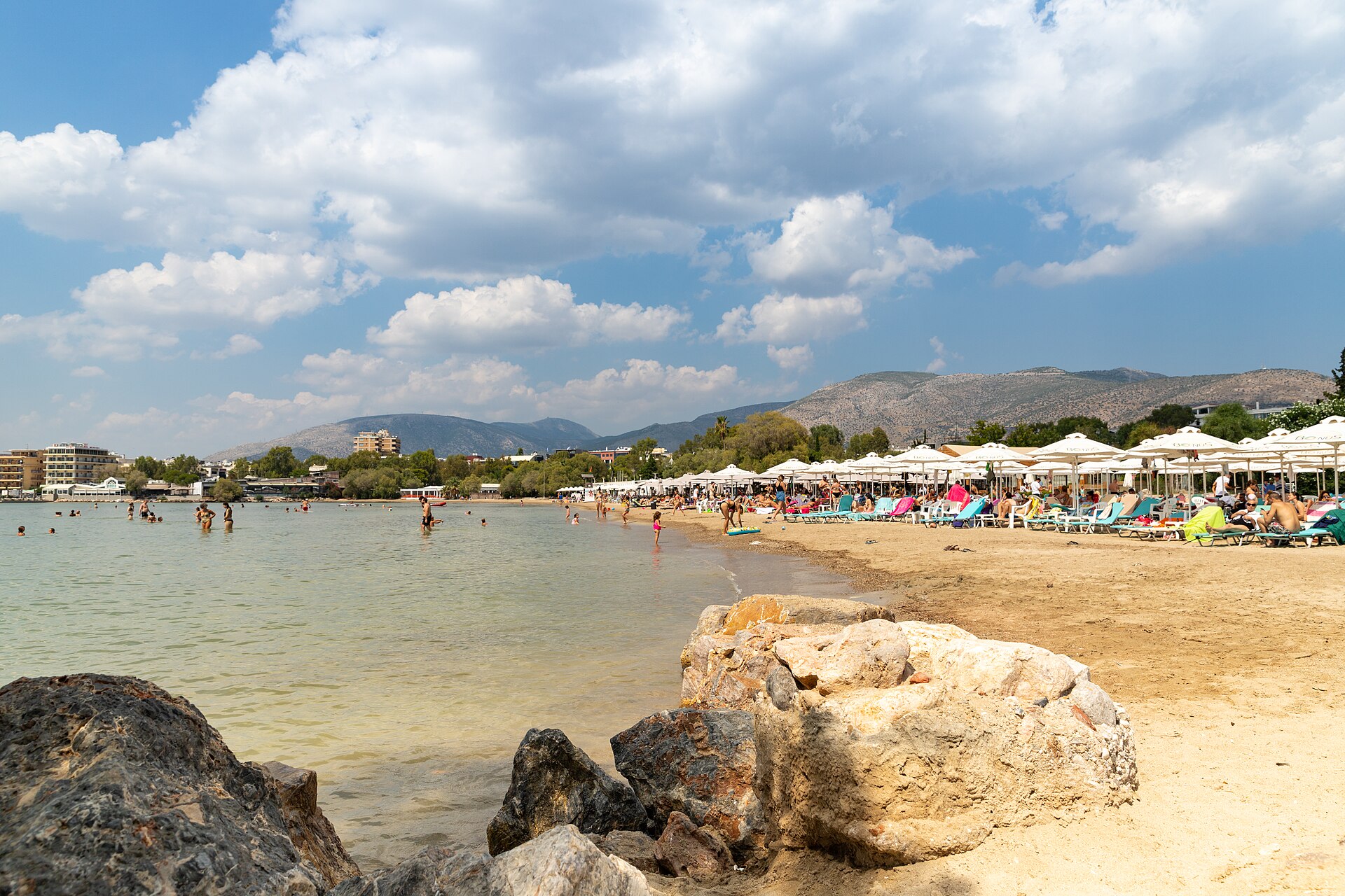 Swimmers wade beside the sandy shore at Asteras beach in Glyfada, Athens, beneath rows of white parasols.
