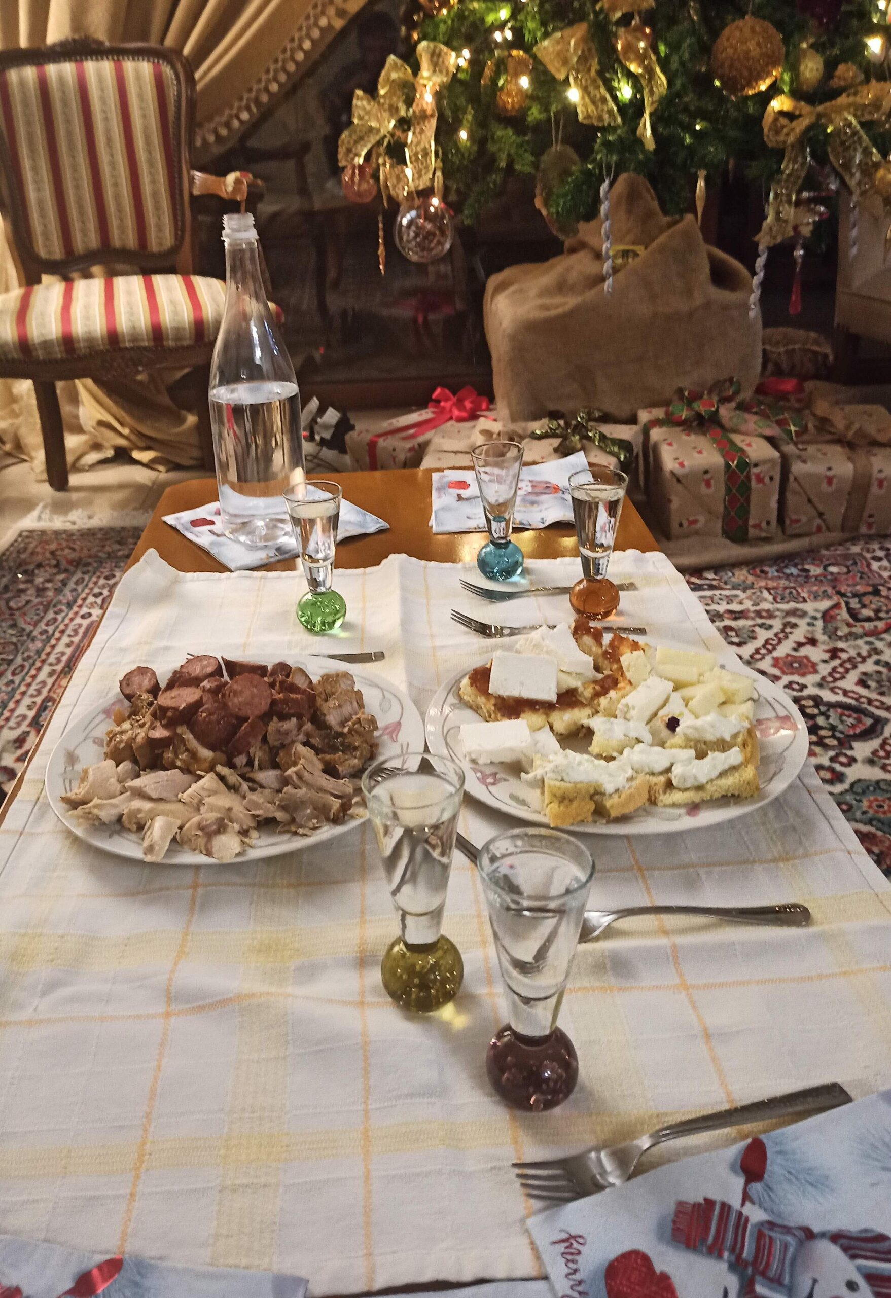 Tsipouro and small stemmed glasses stand beside plates of meat and cheese-topped bread, with a decorated Christmas tree behind.
