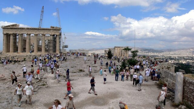 Visitors cross the rocky Acropolis plateau beside the Parthenon, with the Erechtheion and Athens beyond