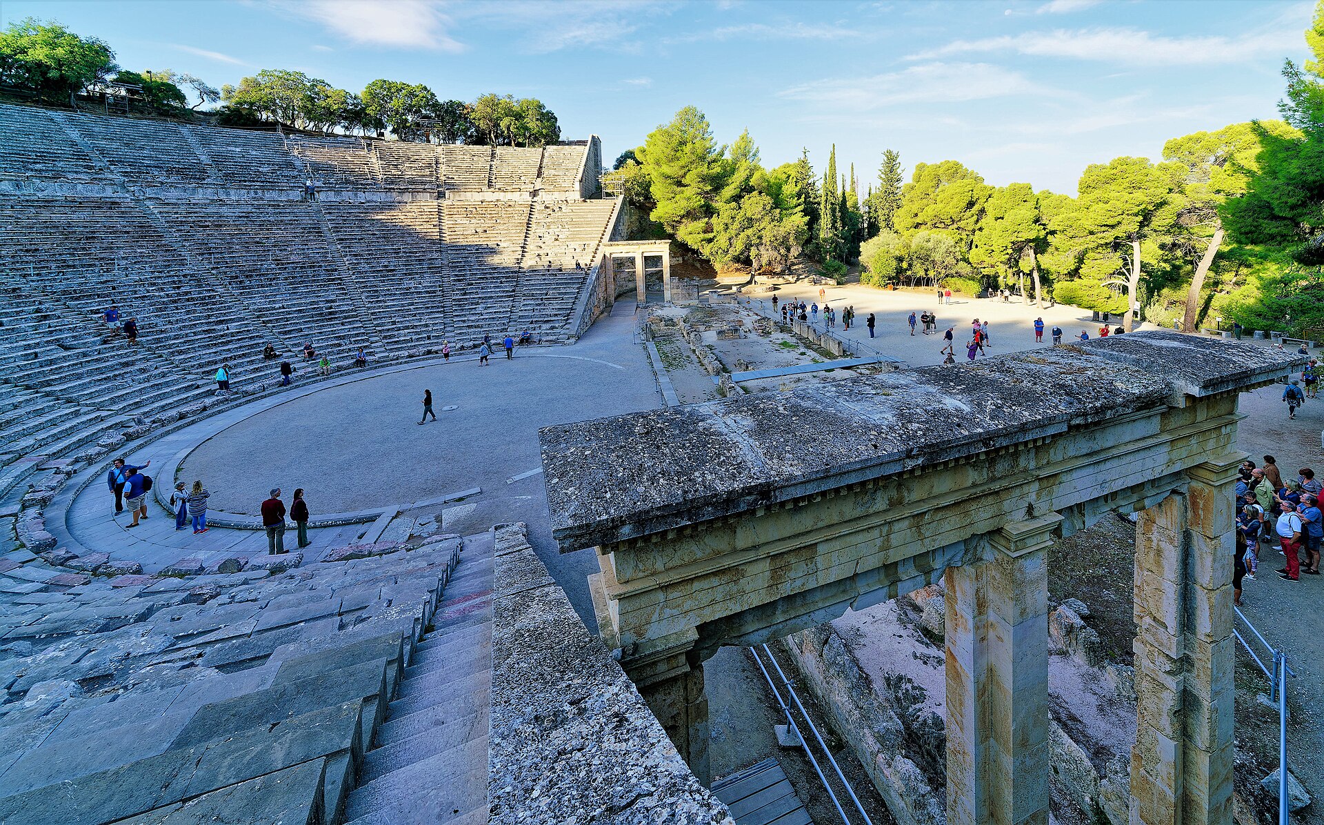Visitors explore the ancient theatre of Epidaurus, where curved stone seating surrounds the orchestra beside a monumental entrance.