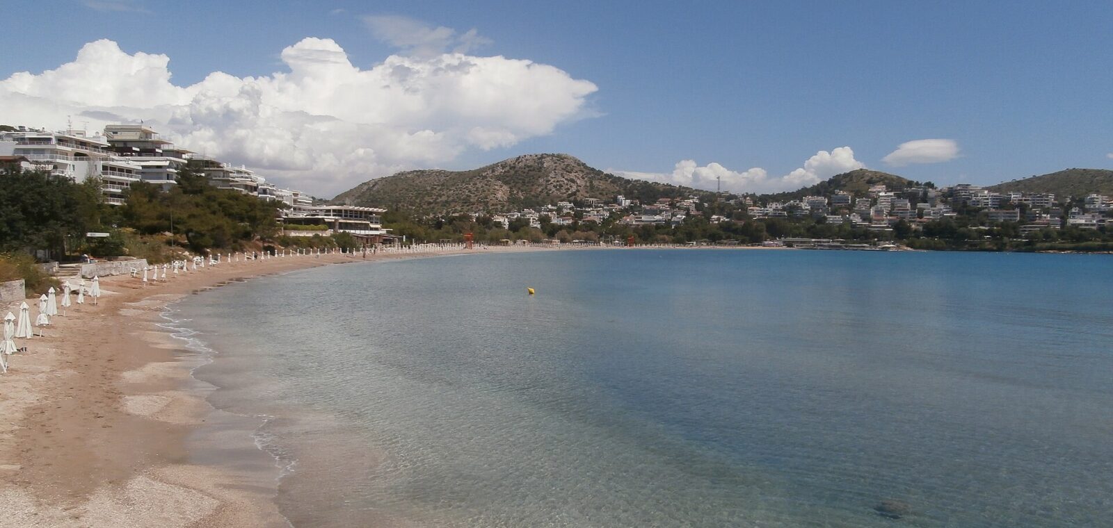 Vouliagmeni beach curves around a clear blue bay, with white parasols and loungers below buildings on the hillside.