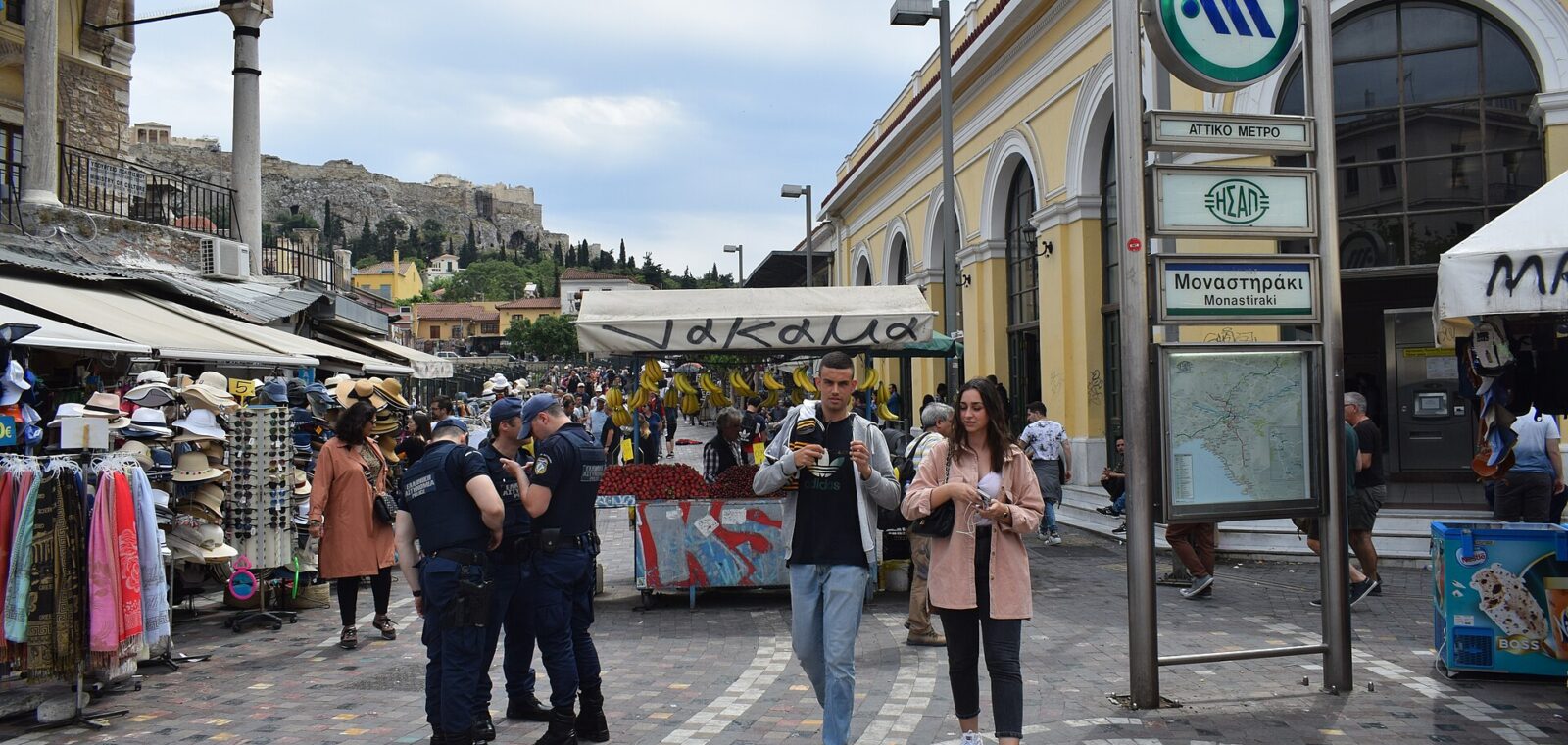 Young adults pass market stalls beside Monastiraki metro station in Athens, with the Acropolis rising in the background.